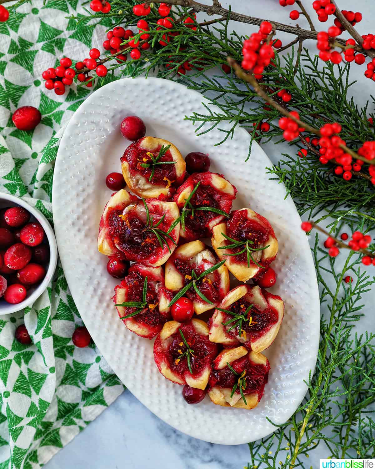 cranberry brie tartlet appetizers on a plate next to plate of pistachio cranberry shortbread cookies and glasses of cranberry mocktails with holiday decor.