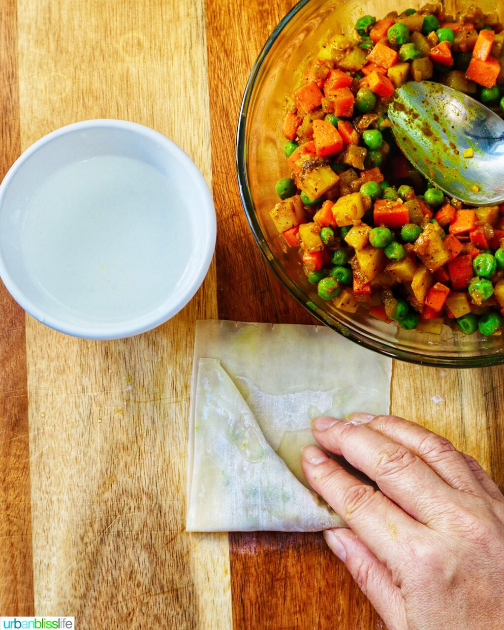 hand folding curry samosa wrapper on a cutting board with bowl of curry samosa filling.