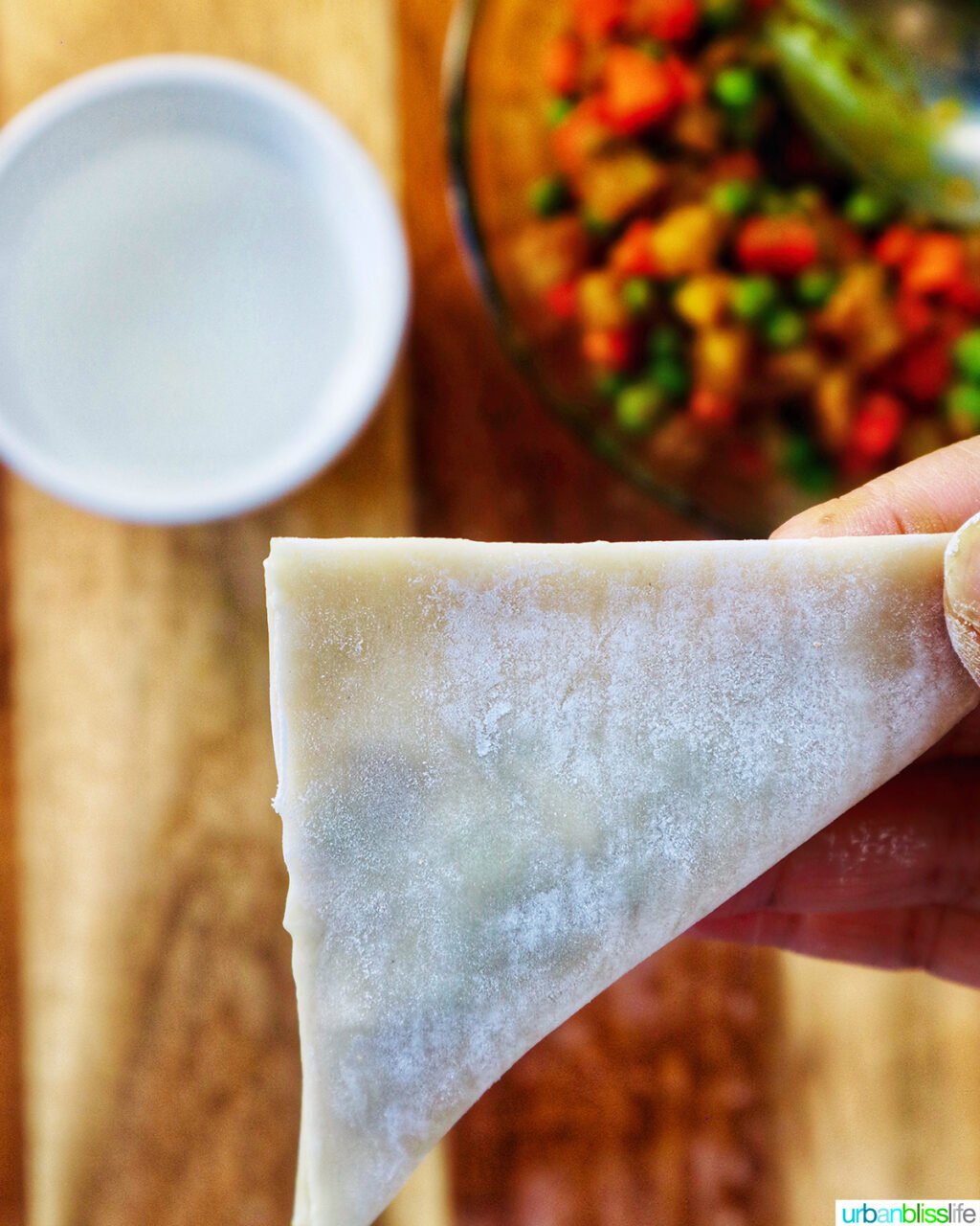 hand folding curry samosa wrapper on a cutting board with bowl of curry samosa filling.