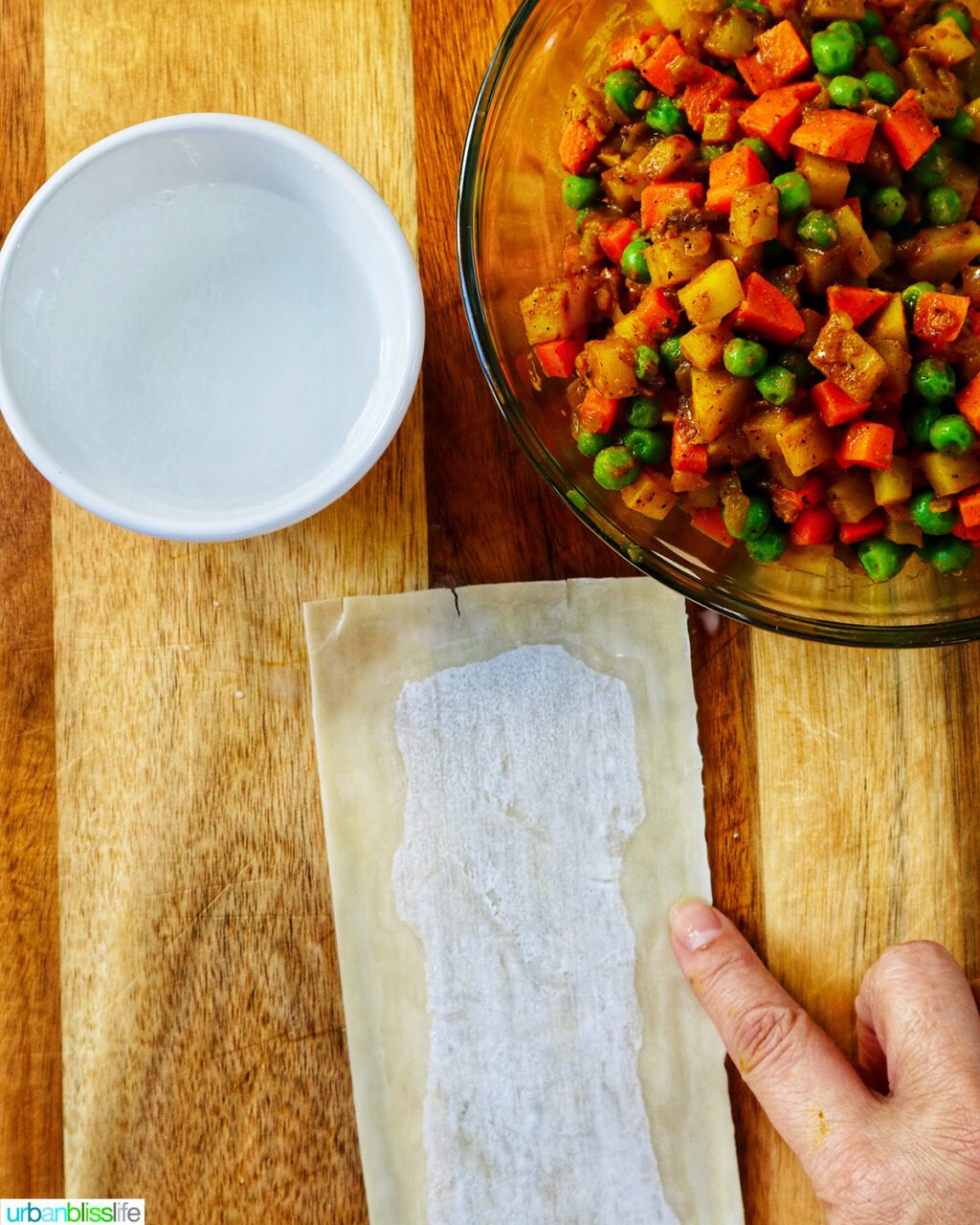 hand wetting the sides of an egg roll wrapper on a cutting board with bowl of curry samosa filling.