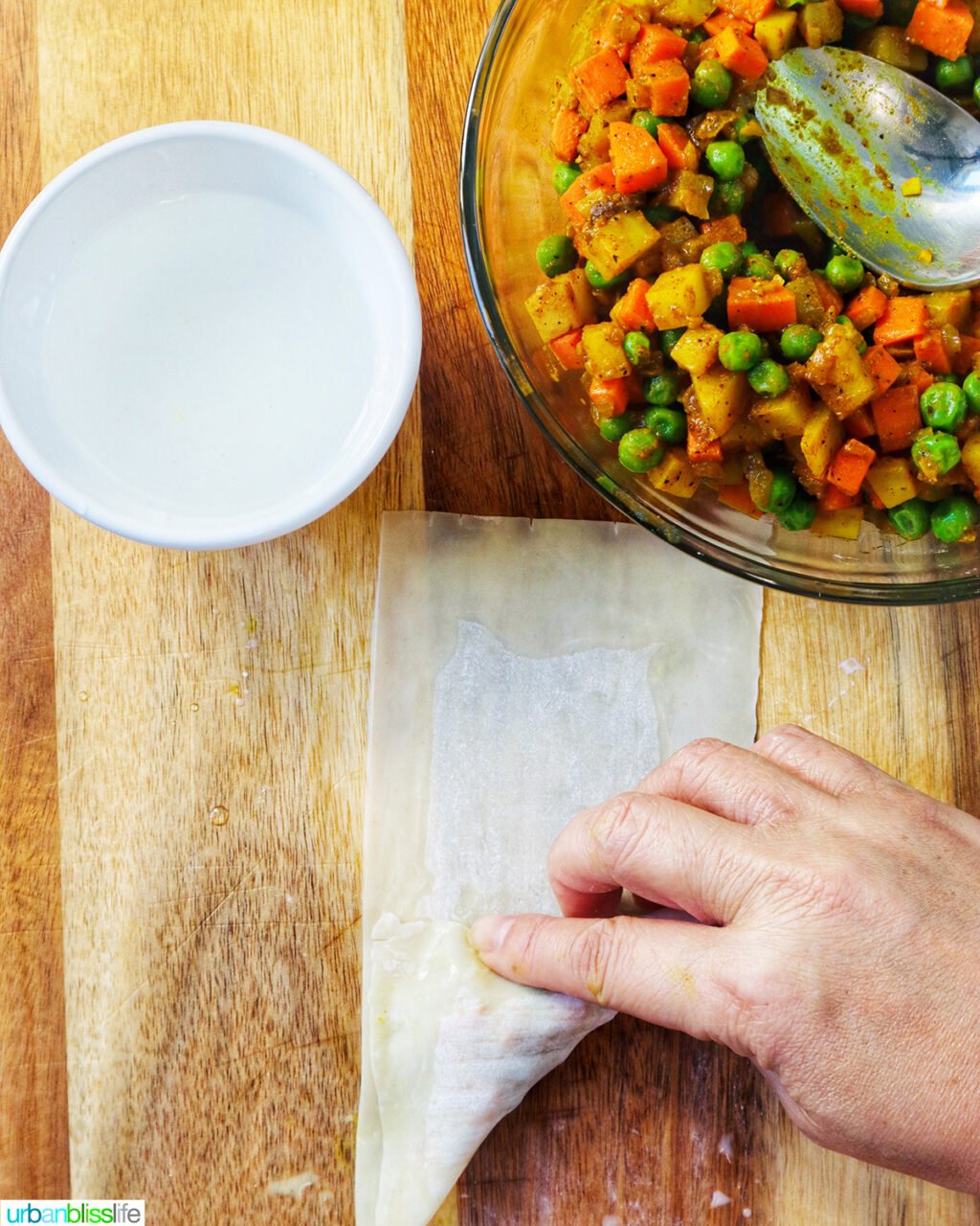 hand folding curry samosa wrapper  on a cutting board with bowl of curry samosa filling.