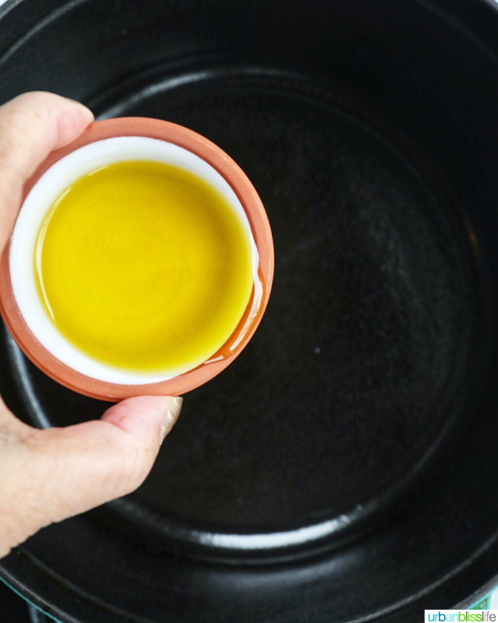 hand holding bowl of olive oil over a dutch oven.