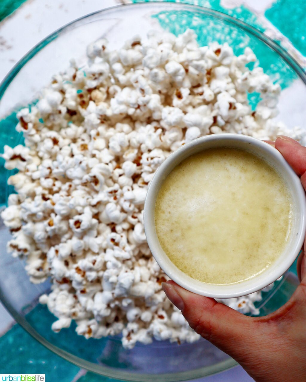 hand holding a bowl of melted butter over a bowl of popcorn.