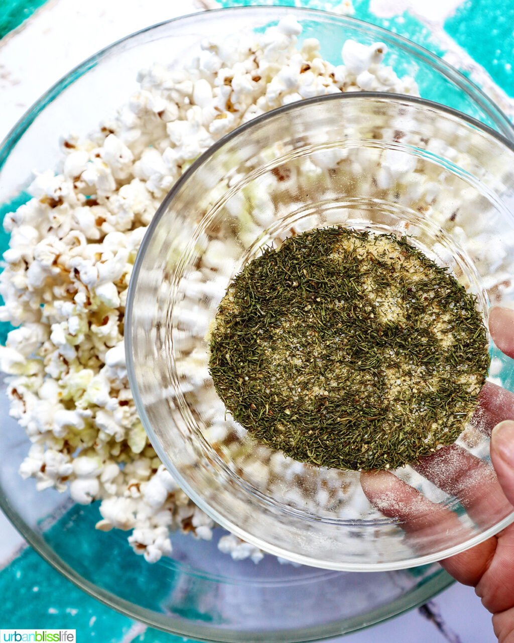 hand holding a bowl of mixed spices and herbs over a bowl of popcorn.
