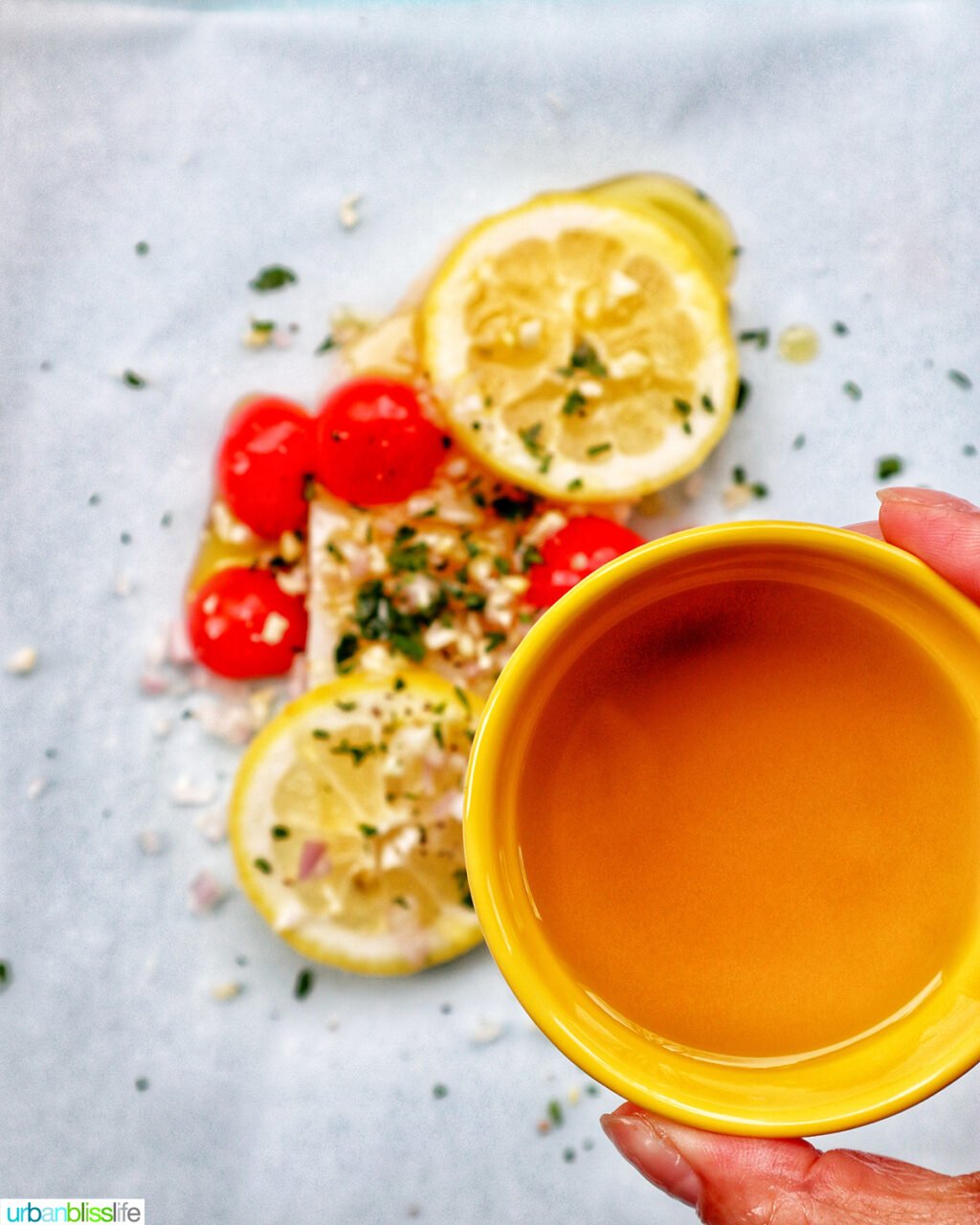 hand holding a bowl of olive oil over fish and veggies.