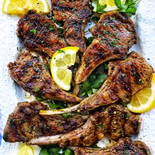 Greek lamb chops on a white plate with herbs and lemon slices.