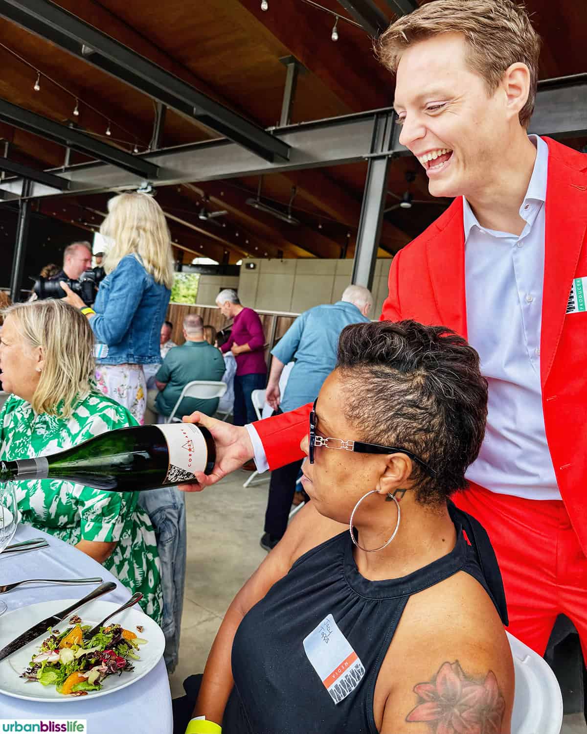 man in red suit pouring sparkling wine into a glass with woman seated at table.