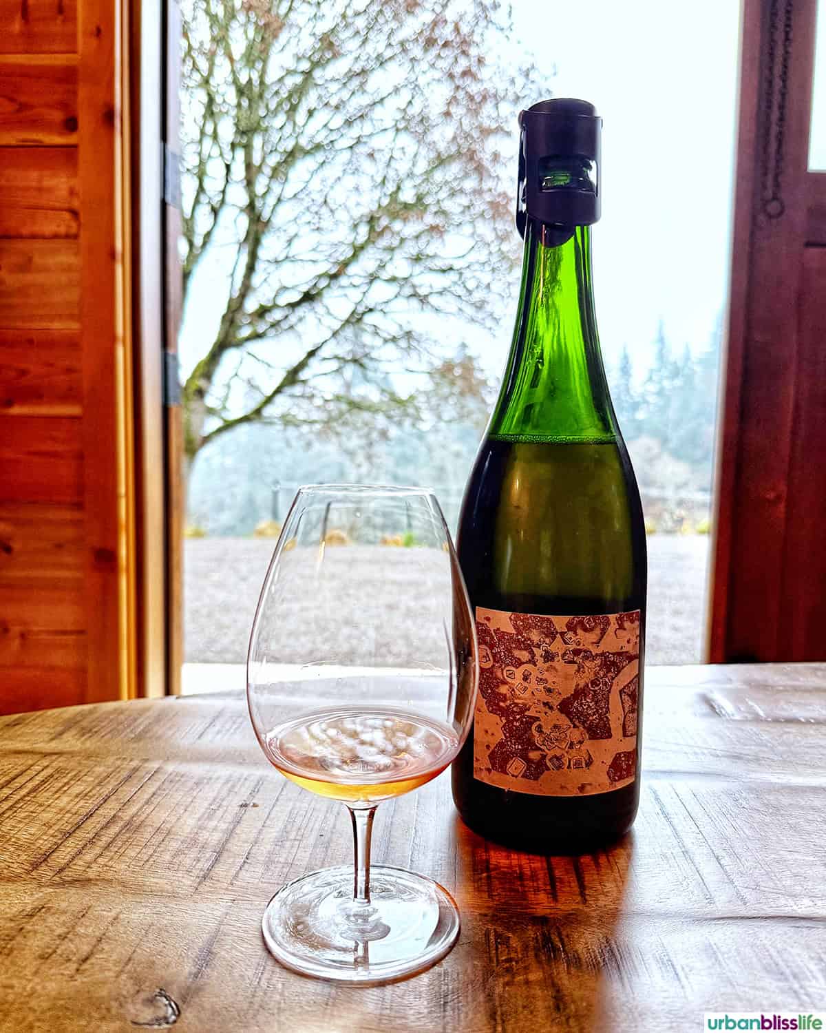 Bottle of Evening Land Vineyards Blanc de Noirs and glass of wine in front of cabin doors leading to the vineyard.