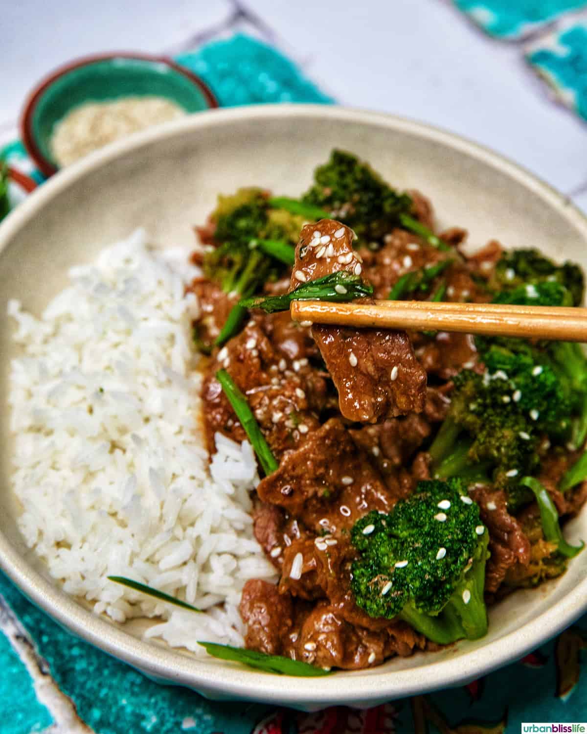 Chopsticks holding up a slice of beef, above slow cooker beef and broccoli with white rice in a bowl.