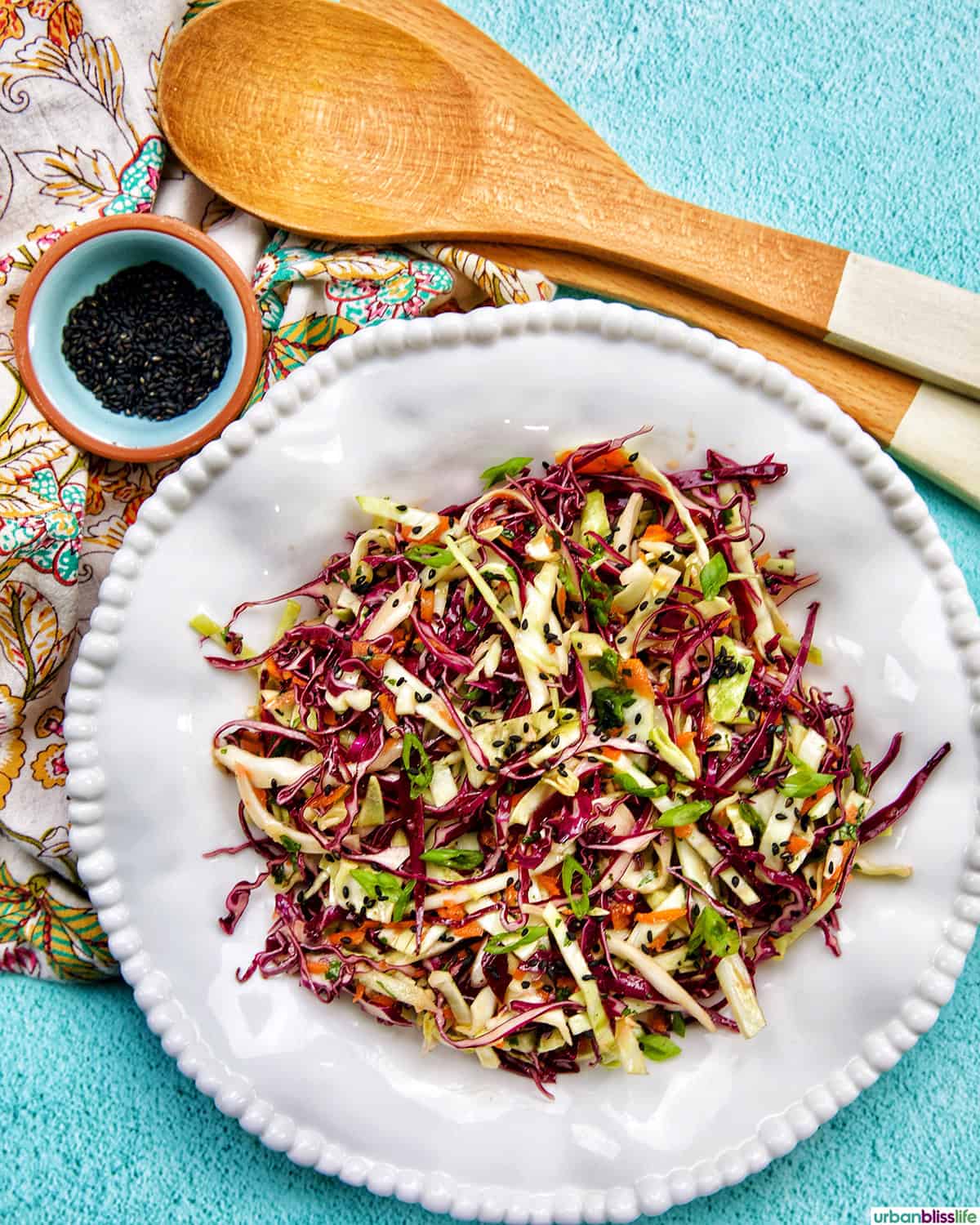 Asian cabbage coleslaw in a white plate with side of black sesame seeds and wooden utensils.