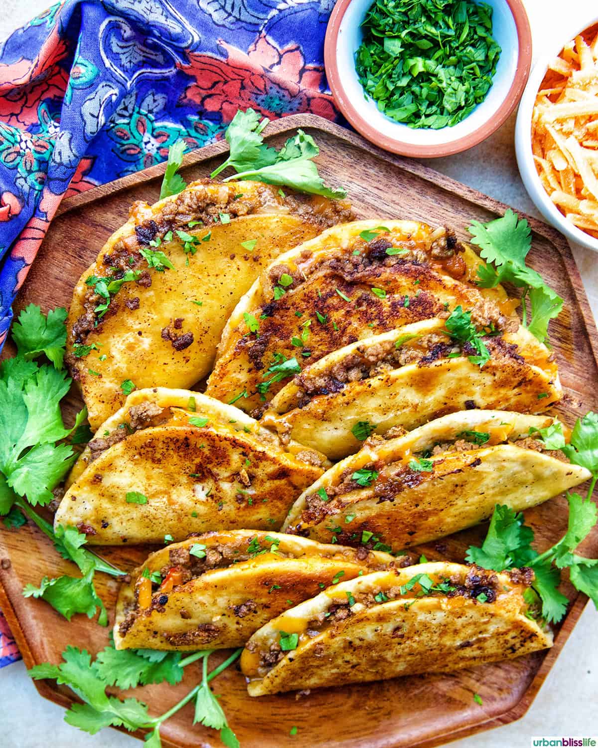plate full of crispy beef tacos with side of chopped cilantro, shredded cheese.