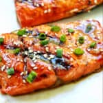 two fillets of BBQ Teriyaki Salmon with green onions and sesame seeds on a white plate.