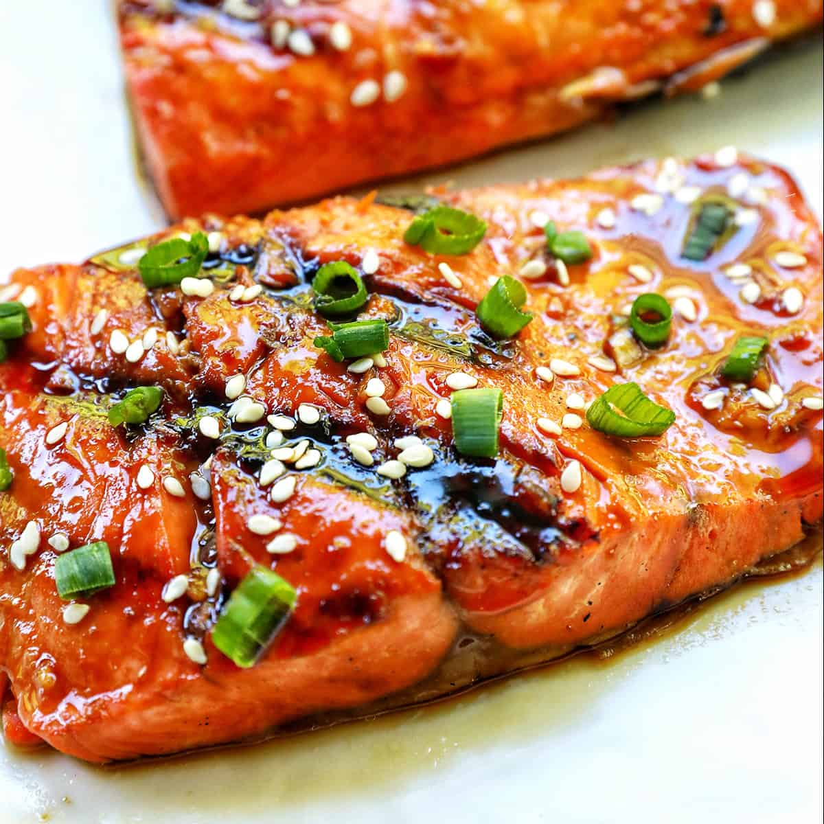 two fillets of BBQ Teriyaki Salmon with green onions and sesame seeds on a white plate.
