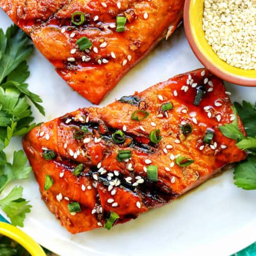 two fillets of BBQ Teriyaki Salmon with green onions and sesame seeds on a white plate.