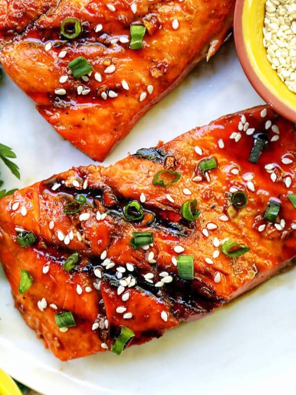 two fillets of BBQ Teriyaki Salmon with green onions and sesame seeds on a white plate.