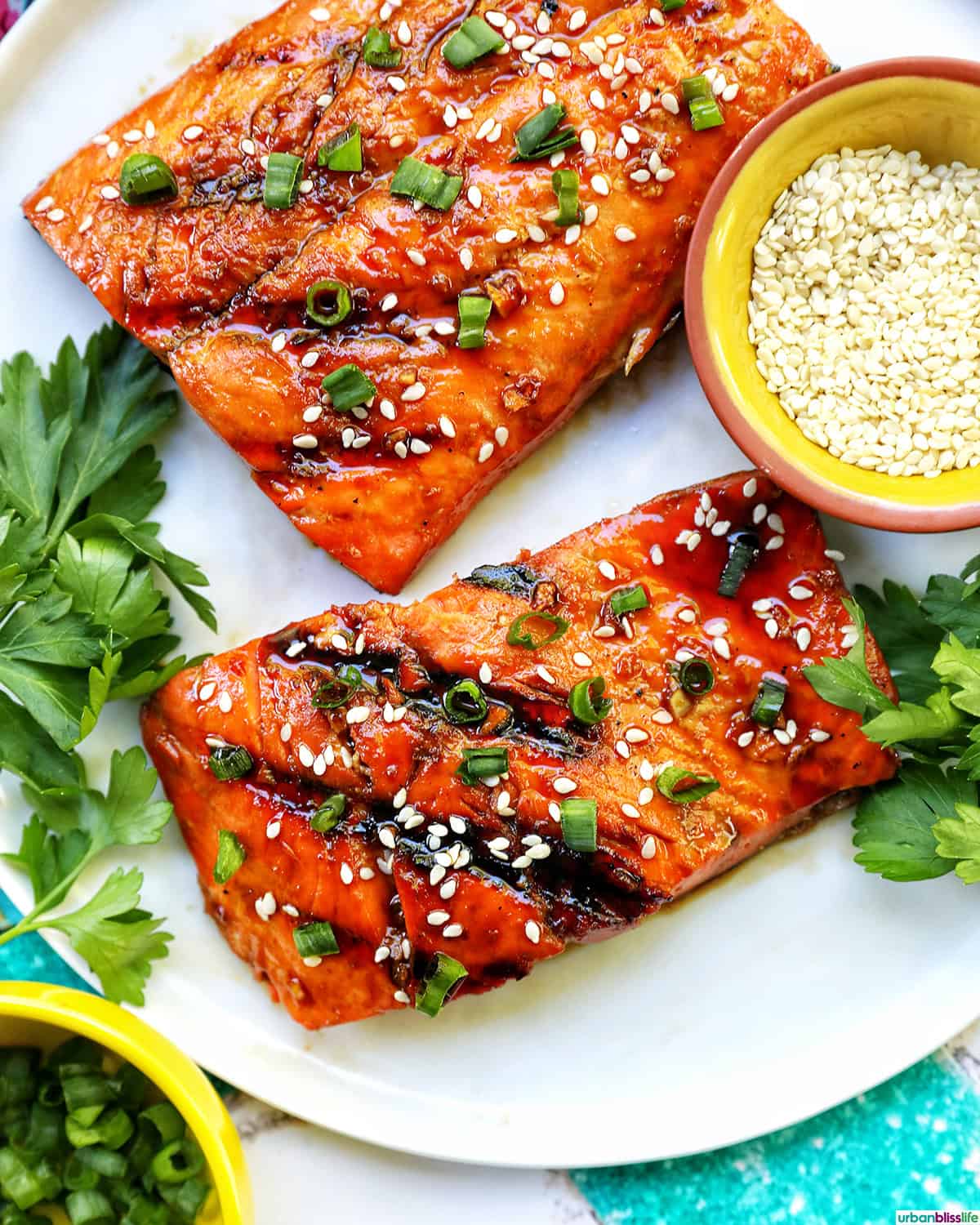 two fillets of BBQ Teriyaki Salmon with green onions and sesame seeds on a white plate.