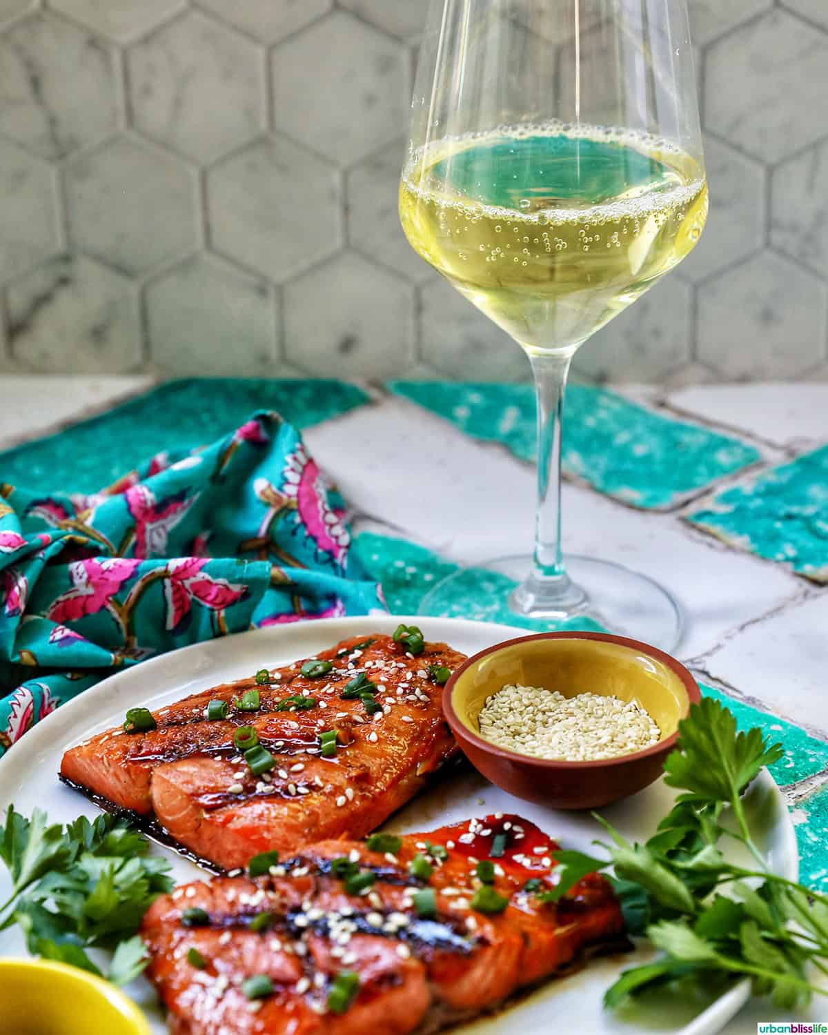 two fillets of BBQ Teriyaki Salmon with green onions and sesame seeds on a white plate and a glass of white wine.