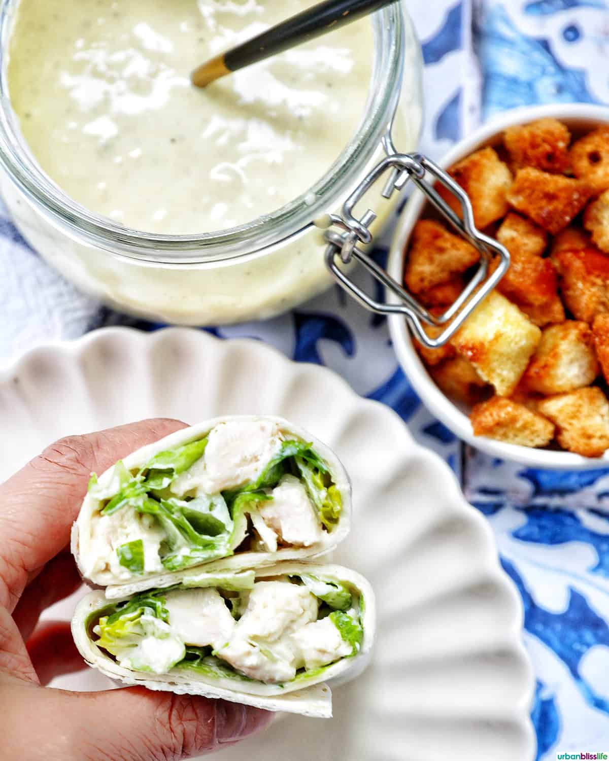 hand holding a Chicken Caesar Salad Wrap sliced in half with side of croutons and dressing.