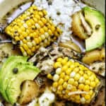 bowl of Colombian Ajiaco soup with corn on the cob and chicken, and sides of rice, avocado slices, capers, and sour cream.