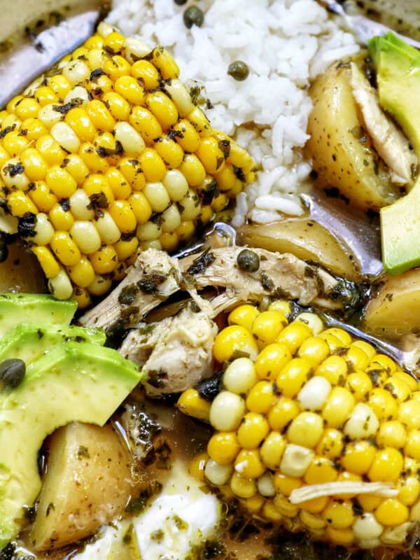 bowl of Colombian Ajiaco soup with corn on the cob and chicken, and sides of rice, avocado slices, capers, and sour cream.