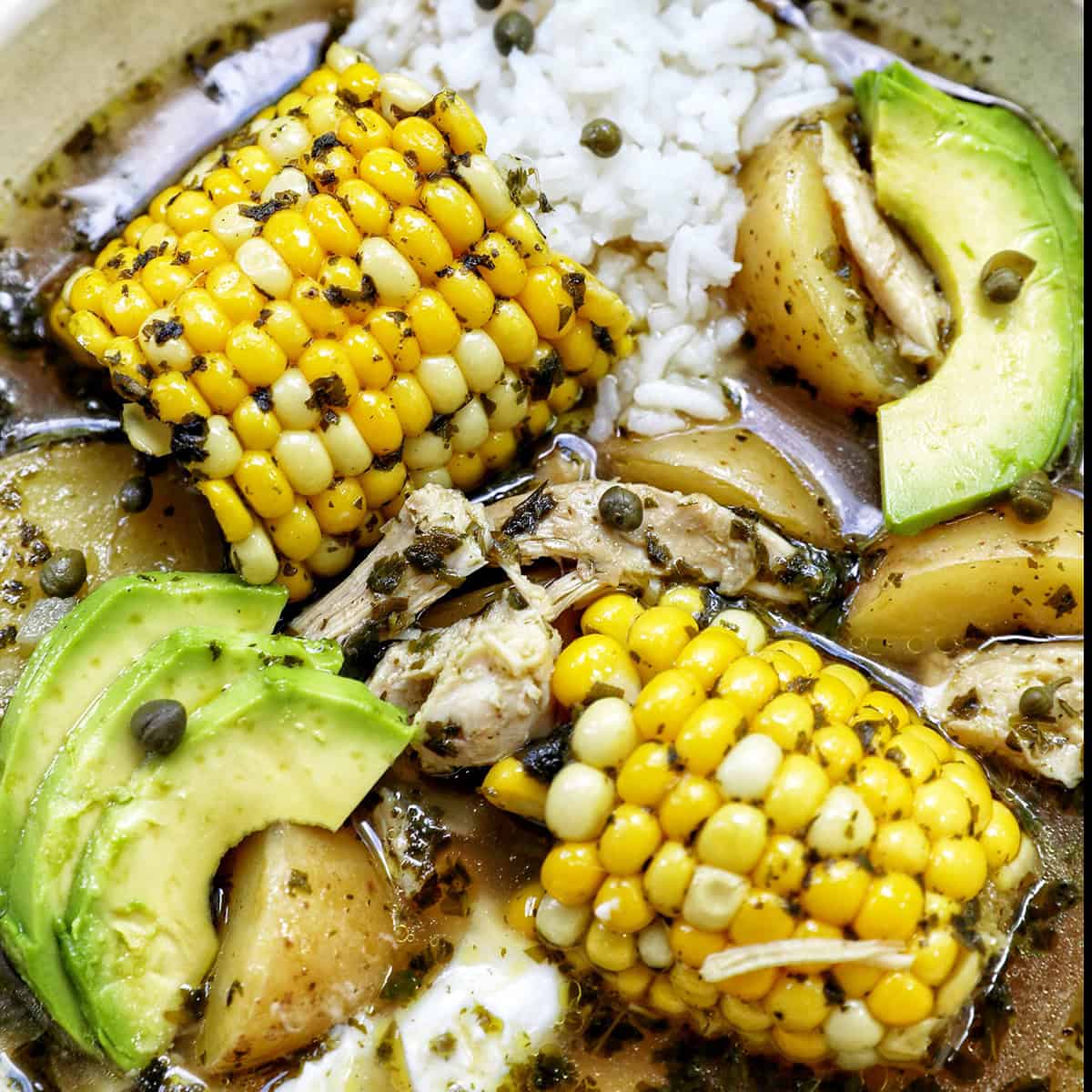 bowl of Colombian Ajiaco soup with corn on the cob and chicken, and sides of rice, avocado slices, capers, and sour cream.