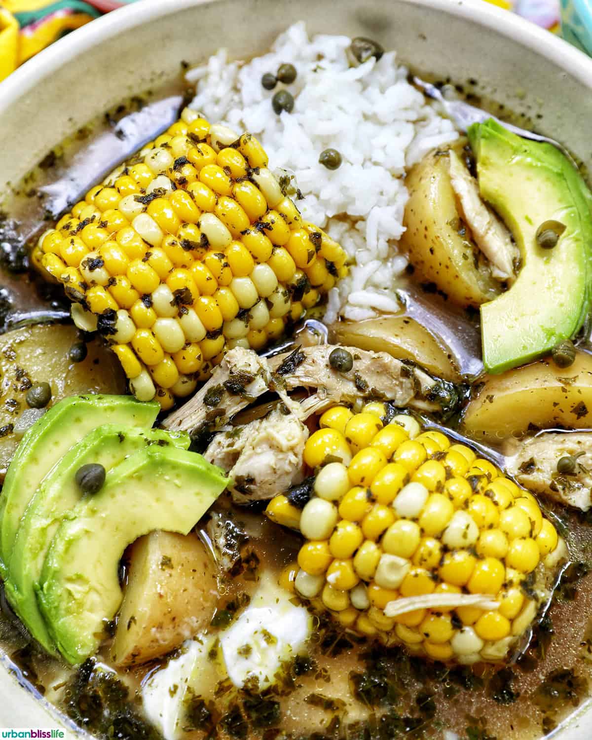 bowl of Colombian Ajiaco soup with corn on the cob and chicken, and sides of rice, avocado slices, capers, and sour cream.