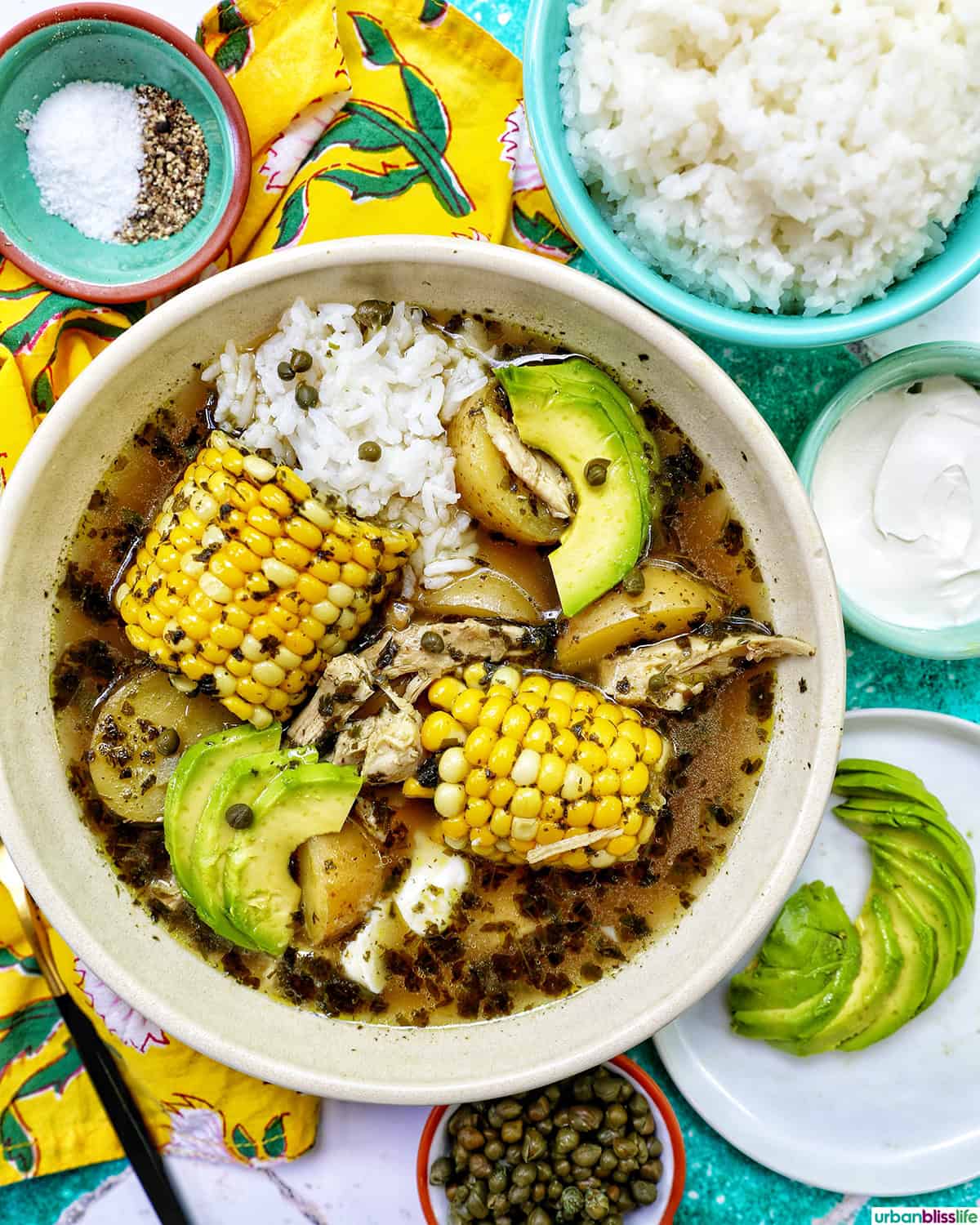 bowl of Colombian Ajiaco soup with corn on the cob and chicken, and sides of rice, avocado slices, capers, and sour cream.
