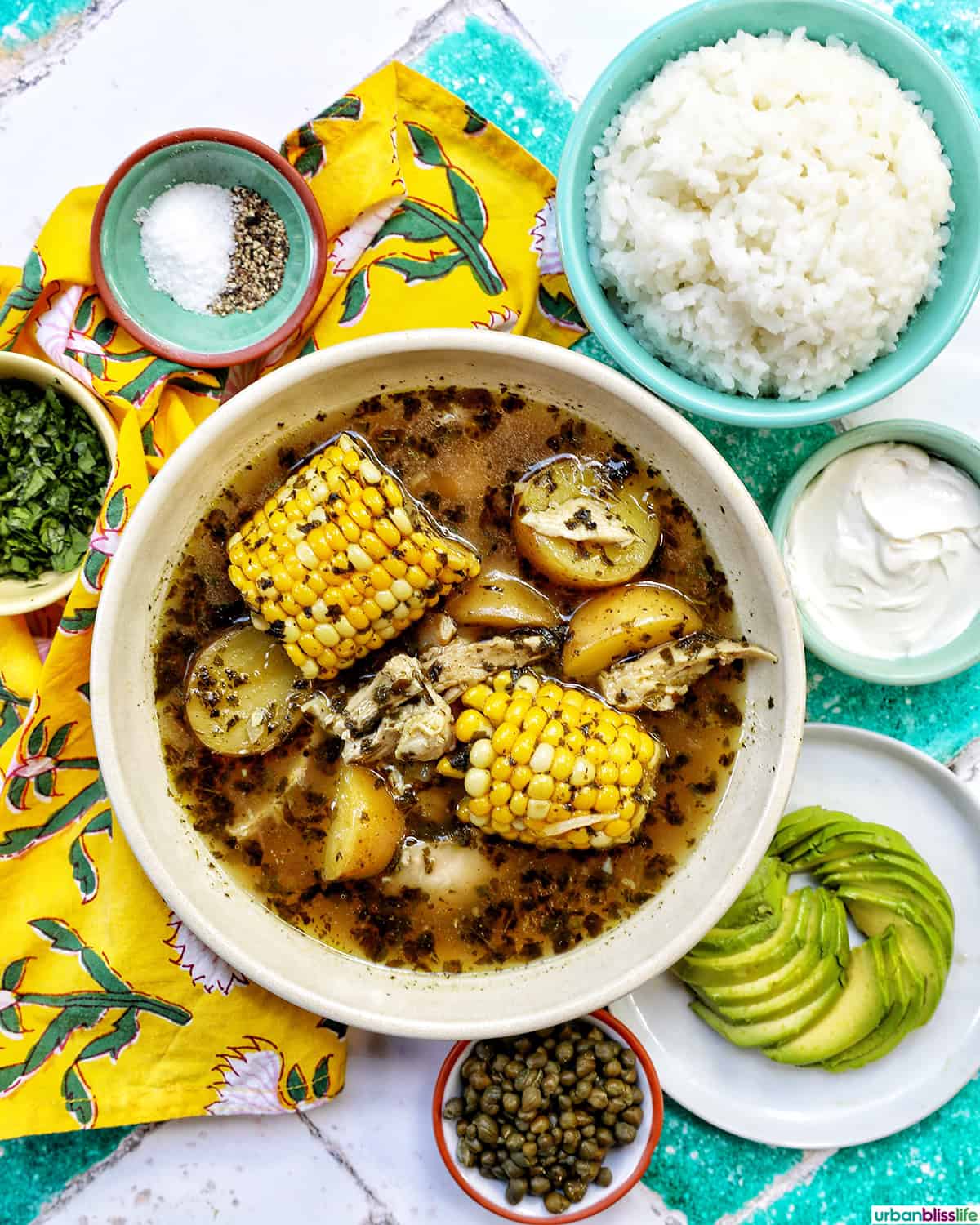 bowl of Colombian Ajiaco soup with corn on the cob and chicken, and sides of rice, avocado slices, capers, and sour cream.