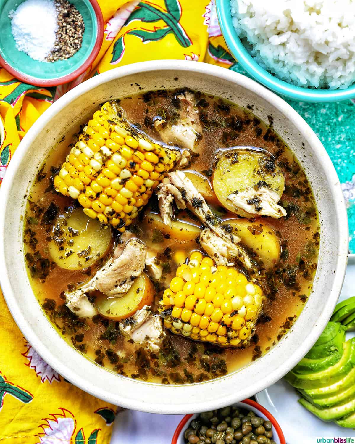 bowl of Colombian Ajiaco soup with corn on the cob and chicken, and sides of rice, avocado slices, capers, and sour cream.