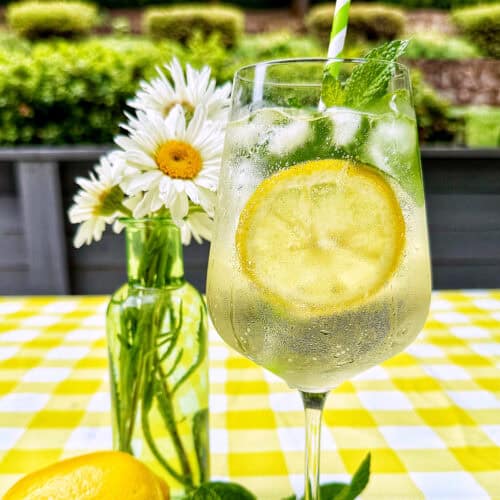Hugo Spritz cocktail surrounded by mint leaves, limes, lemons, and vase of daisy flowers on yellow checkered tablecloth.