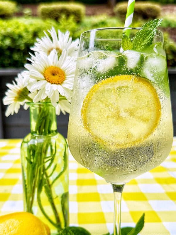 Hugo Spritz cocktail surrounded by mint leaves, limes, lemons, and vase of daisy flowers on yellow checkered tablecloth.
