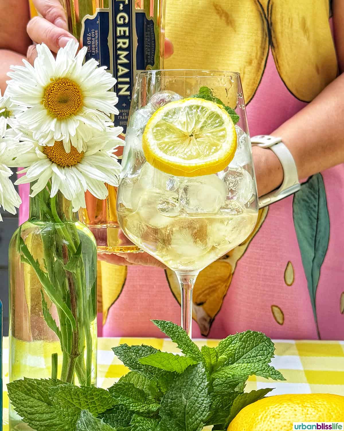 Hugo Spritz cocktail surrounded by mint leaves, limes, lemons, and vase of daisy flowers on yellow checkered tablecloth.