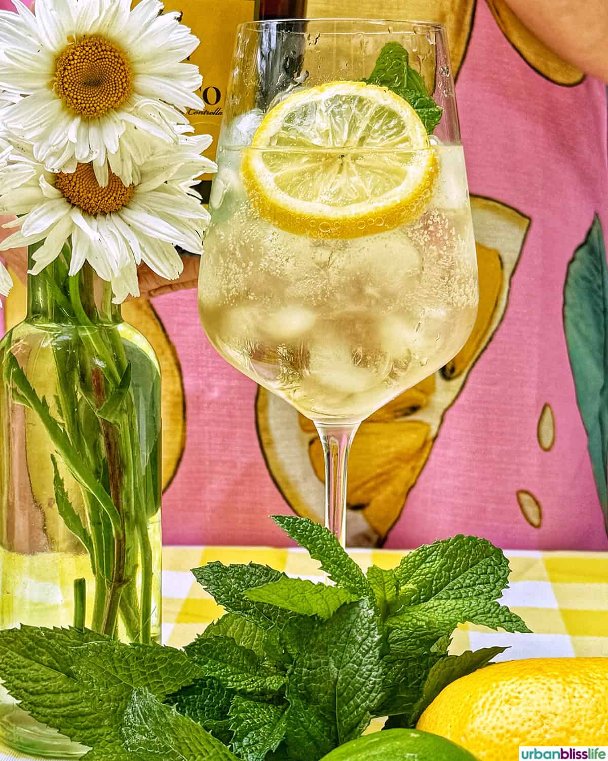 Hugo Spritz cocktail surrounded by mint leaves, limes, lemons, and vase of daisy flowers on yellow checkered tablecloth.