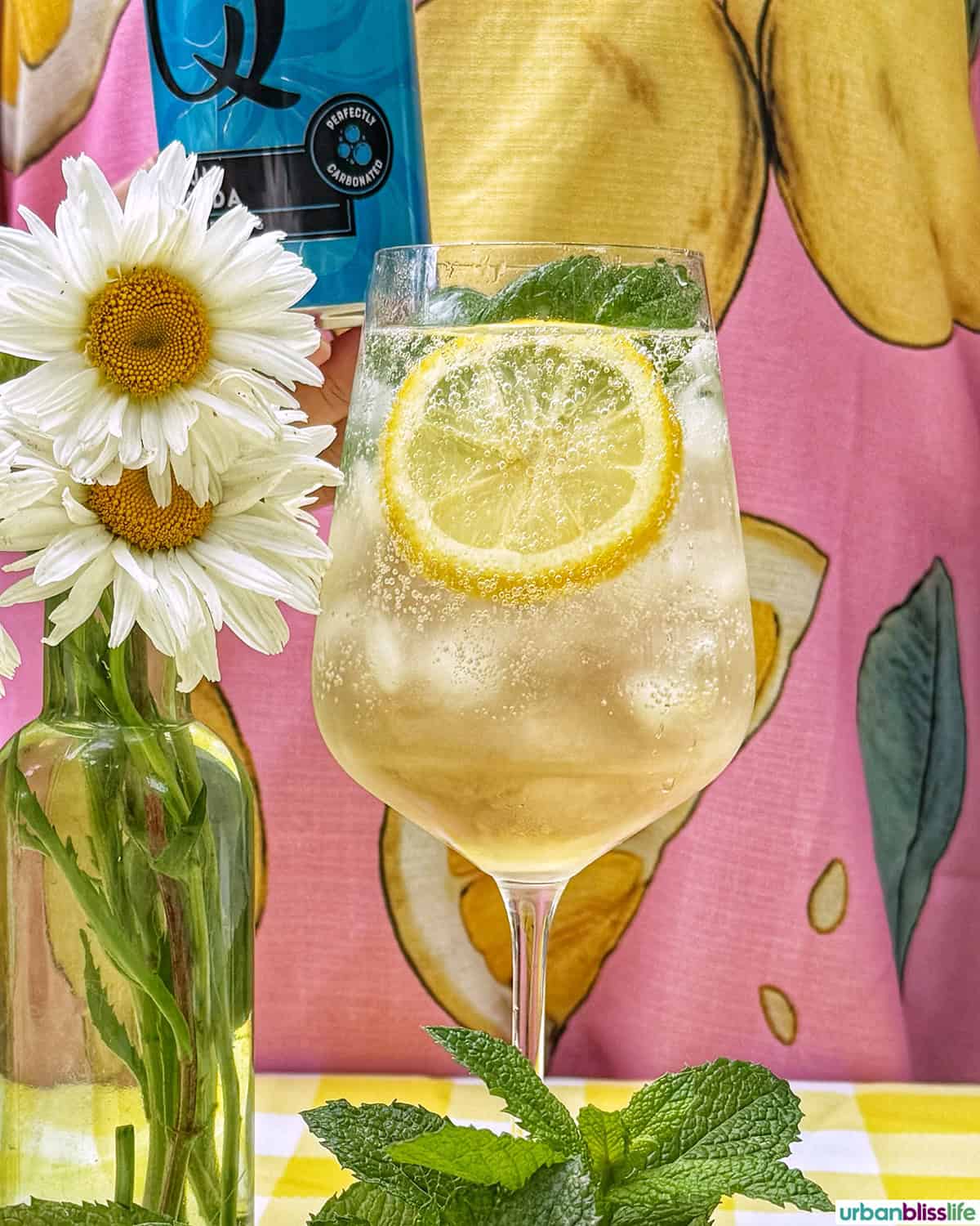 Hugo Spritz cocktail surrounded by mint leaves, limes, lemons, and vase of daisy flowers on yellow checkered tablecloth.