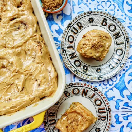 baking dish with coffee cinnamon rolls against a blue and white French backdrop and two plates with cinnamon rolls on them.