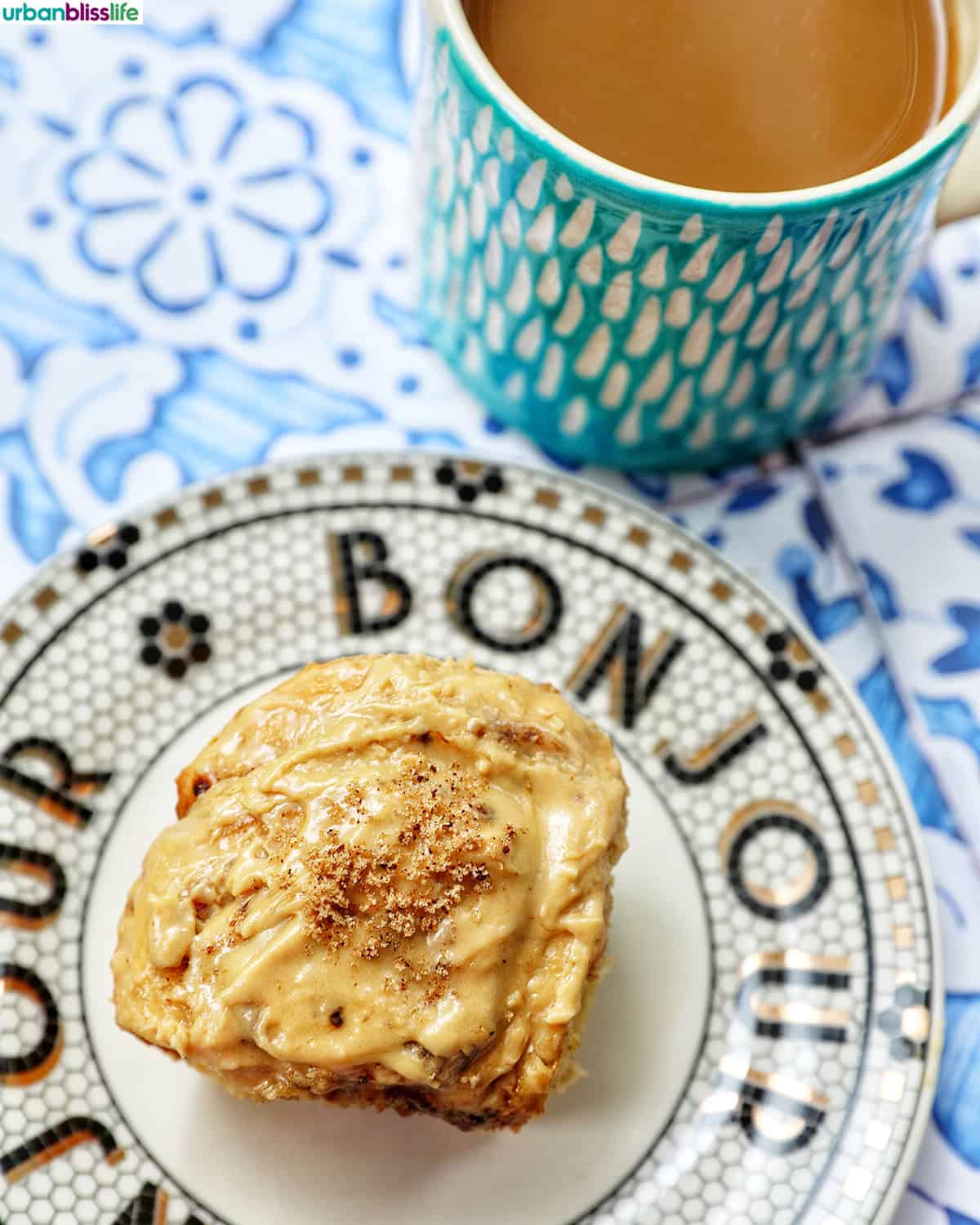Bonjour plate with a coffee cinnamon roll on it next to a cup of coffee.