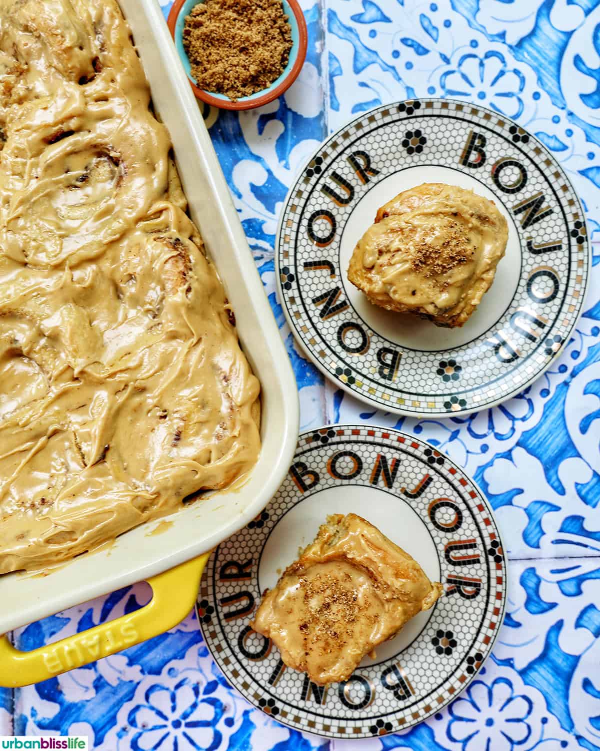 baking dish with coffee cinnamon rolls against a blue and white French backdrop and two plates with cinnamon rolls on them.